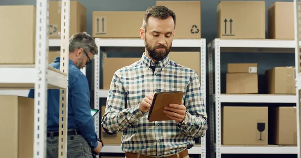 邮递员站在邮局里 手里拿着包裹 在平板电脑上窃听器 男工带着电脑上网 在满箱子的产房里登记 — 图库照片