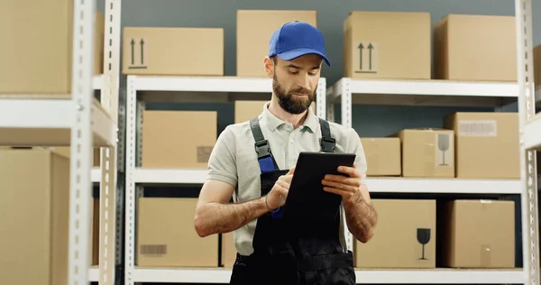 Portrait of delivery man in postal store with parcels and tapping on tablet computer. Male worker with digital tablet device doing registration in delivery room full of boxes.