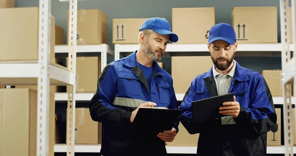 Portrait of two delivery man in blue uniforms standing in postal store and talking while working. Men of delivery work with folder of documents and tablet device discussing parcels.