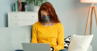 Maskeli neşeli kızıl saçlı kız evdeki oturma odasında koltukta oturuyor ve gümüş dizüstü bilgisayar kullanıyor..