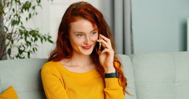Mutlu ve pozitif bir kız evinde koltukta oturmuş akıllı telefonuyla konuşuyor. İletişim uygulaması akıllı telefon kavramı