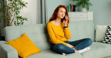 Mutlu ve pozitif bir kız evinde koltukta oturmuş akıllı telefonuyla konuşuyor. İletişim uygulaması akıllı telefon kavramı