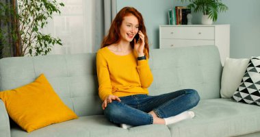 Güzel, neşeli, kızıl saçlı bir kızın portresi. Rahat ol, koltukta otur ve cep telefonunu kullan..