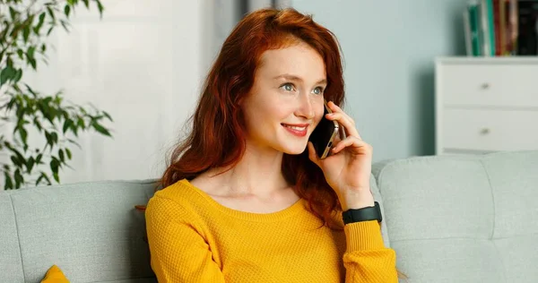 Счастливая и позитивная девушка сидит дома на диване и разговаривает по смартфону. Концепция смартфона — стоковое фото