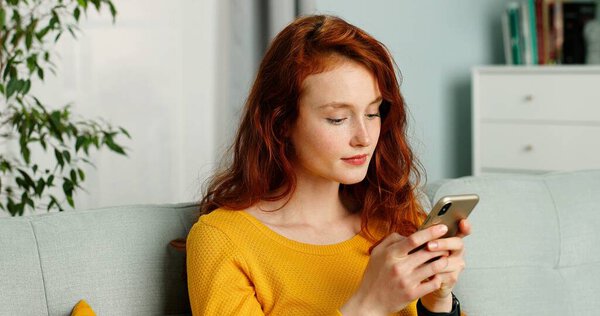 Красивая молодая рыжая женщина с кудрявыми волосами пишет смс на смартфоне дома
.