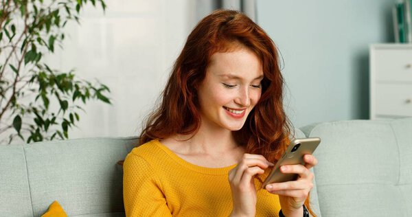 Счастливая и позитивная девочка сидит дома на диване и пользуется смартфоном
.