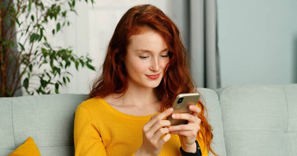 Красивая молодая рыжая женщина с кудрявыми волосами пишет смс на смартфоне дома
.