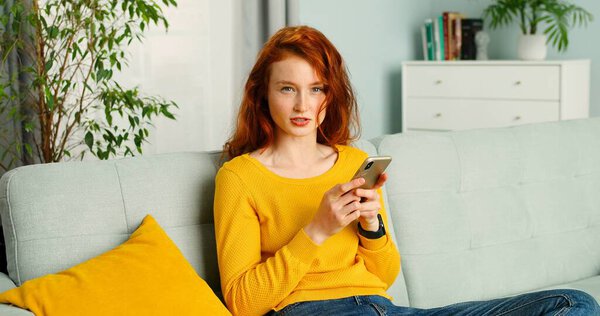 Счастливая и позитивная девочка сидит дома на диване и пользуется смартфоном
.