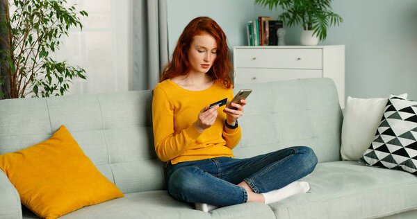 Молодая красивая рыжеволосая женщина сидит на диване в гостиной и делает покупки онлайн на мобильном телефоне
.