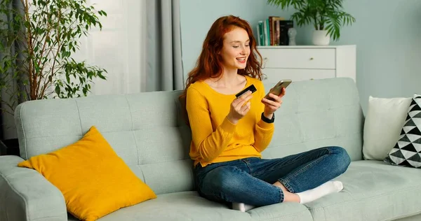 Young beautiful red-haired woman sitting on couch in living room and shopping online on mobile phone. — Stock Photo, Image