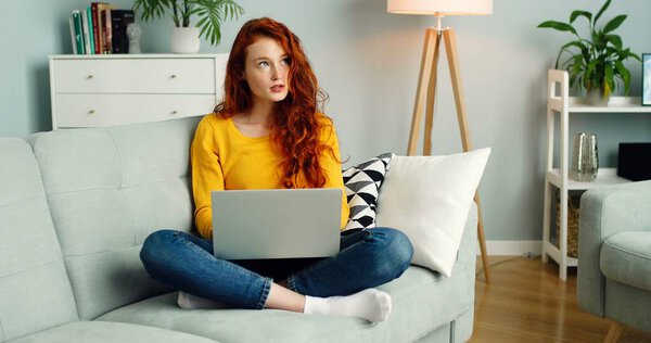 Beautiful cheerful redhead girl using silver laptop while sitting on sofa in living room at home. — Stock Photo, Image
