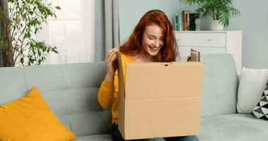 Oturma odasında kanepede oturan ve karton kutuyu açan genç ve güzel bir kadın. Neşeli kız evden paket alıyor. Eve posta teslimatı.