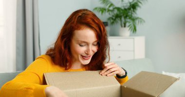 Oturma odasında kanepede oturan ve karton kutuyu açan genç ve güzel bir kadın. Neşeli kız evden paket alıyor. Eve posta teslimatı.