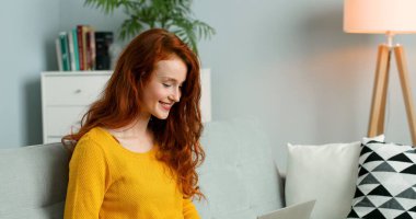 Genç, güzel, kızıl saçlı bir kadın oturma odasında kanepede oturuyor ve internetten bilgisayar kullanarak alışveriş yapıyor..
