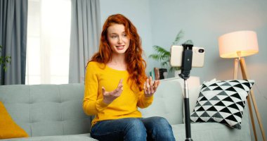 Portrait of young female blogger sitting on couch and talking in front of smartphone webcam. Girl coaching for her vlog online from home. Attractive happy woman coach talk for videoblog. clipart