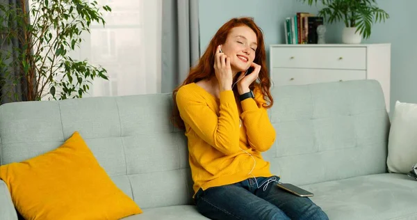 Молодая красивая девушка в наушниках сидит дома на диване и слушает музыку на смартфоне, поет и улыбается . — стоковое фото