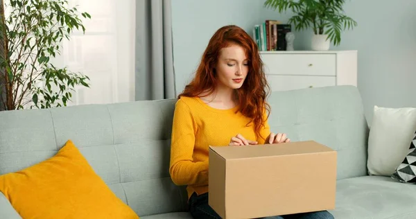 Mujer bonita joven sentada en el sofá en la sala de estar y abrir la caja de cartón. Chica alegre recibiendo paquete en casa. Entrega de correo a domicilio . —  Fotos de Stock