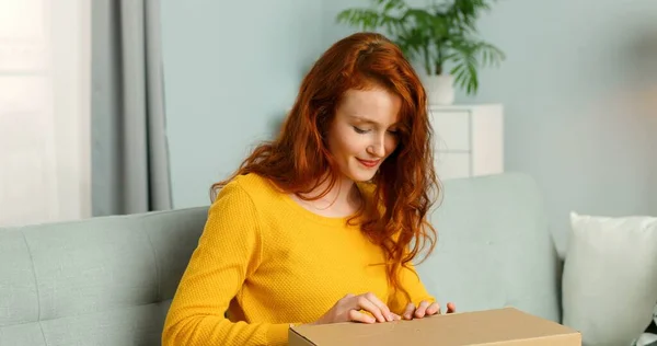 Mujer bonita joven sentada en el sofá en la sala de estar y abrir la caja de cartón. Chica alegre recibiendo paquete en casa. Entrega de correo a domicilio . —  Fotos de Stock