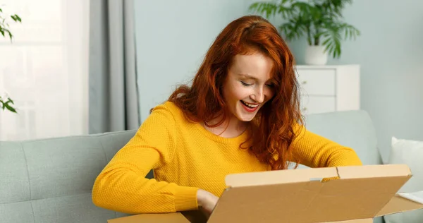 Joven chica alegre recibiendo paquete en casa. Linda chica sentada en el sofá en casa y abriendo la caja de cartón. Entrega de correo a domicilio . —  Fotos de Stock