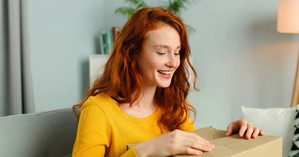 Young cheerful girl getting parcel at home. Pretty girl sitting on sofa at home and opening carton box. Mail delivery to house. — Stock Photo, Image