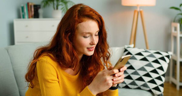 Счастливая и позитивная девочка сидит дома на диване и пользуется смартфоном
.