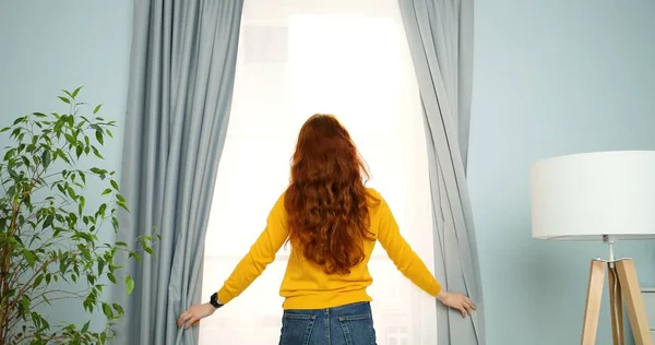 Traseira de mulher jovem com longos cabelos encaracolados vermelhos em pé na sala de estar na janela e cortinas de abertura pela manhã. Visão traseira . — Fotografia de Stock