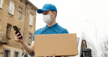 Maskeli genç bir kuryenin portresi arabanın yanında elinde akıllı telefon kullanarak duruyor..