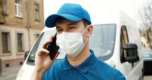 스마트폰을 사용하여 상자를 들고 차 옆에서 서 마스크를 쓴 젊은 택배의 모습. — 스톡 사진