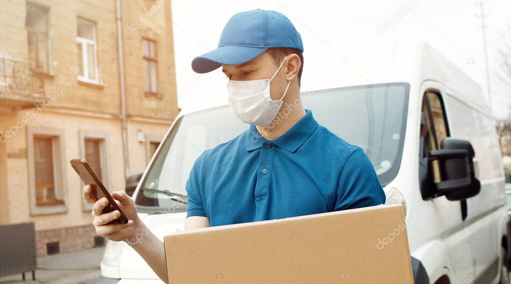 Portrait of young courier in mask standing near car holding carton box using smartphone.