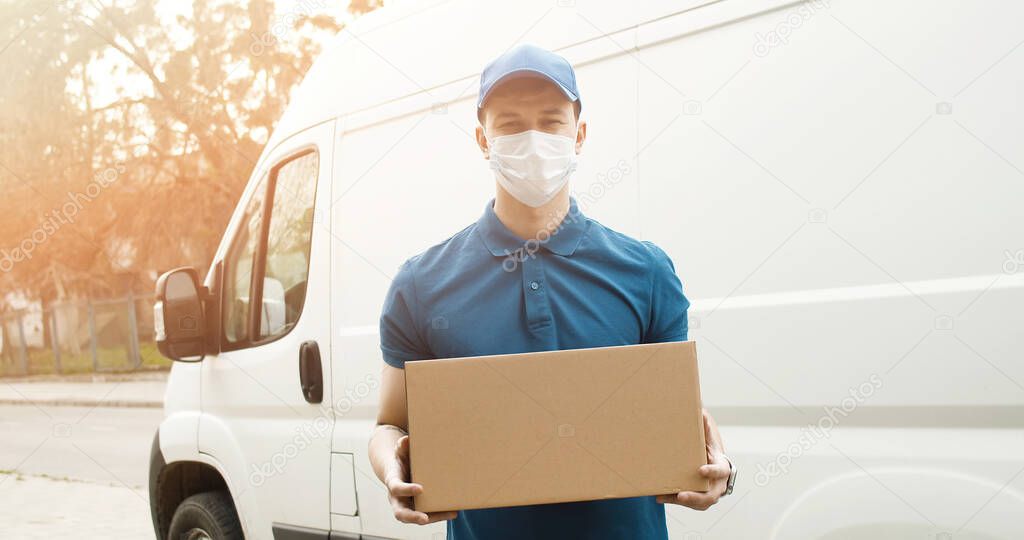 Portrait of young courier in mask standing near delivery car and holding carton box