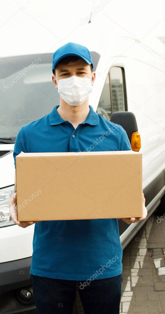 Portrait of young courier in mask standing near delivery car and holding carton box