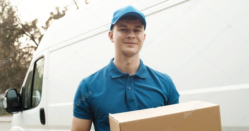 Portrait of young courier in mask standing near delivery car and holding carton box
