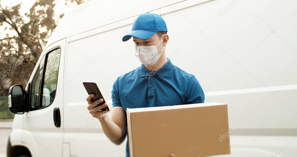 Portrait of young courier in mask standing near car holding carton box using smartphone.