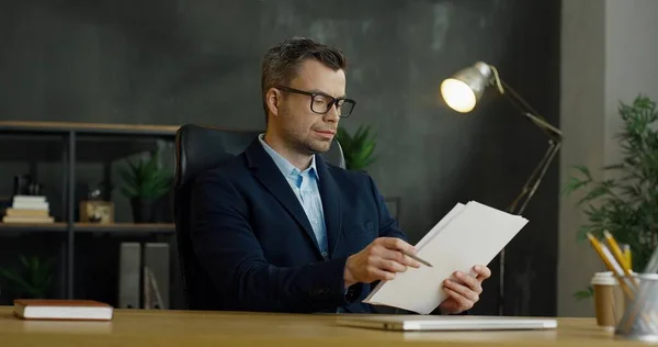 Snygg vit affärsman i glasögon som sitter vid bordet på kontoret och kollar dokument med penna i händerna. — Stockfoto