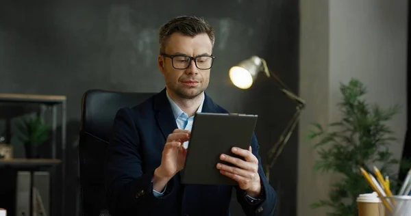 Caucasian businessman in glasses holding and using tablet device in hands. Male texting and typing on screen and thinking in cabinet. — Stock Photo, Image