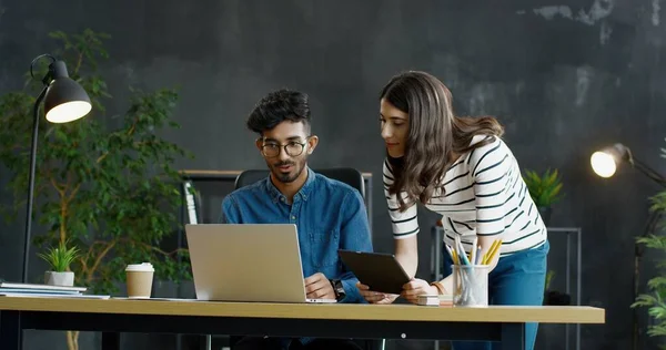 Arabiska killen sitter vid skrivbordet med bärbar dator och vit tjej står bredvid honom med surfplatta enhet. — Stockfoto