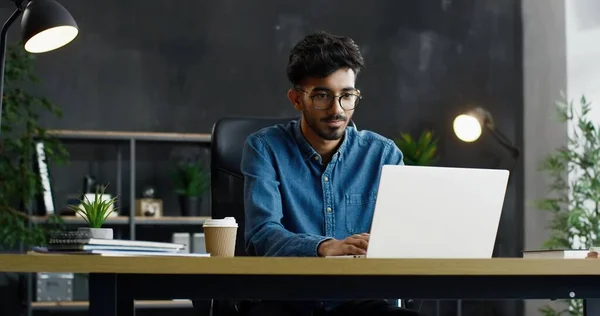 Snygg ung arabisk upptagen manlig kontorsarbetare sitter vid bordet och arbetar på bärbar dator. — Stockfoto