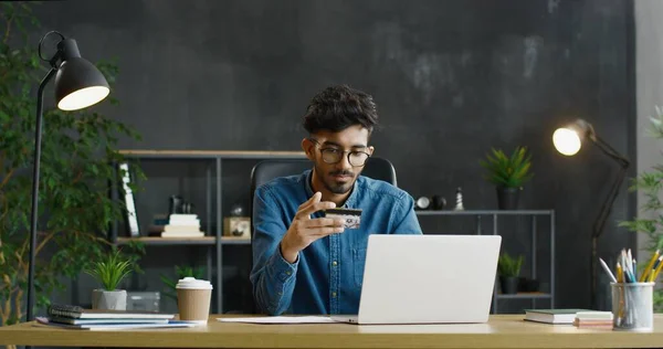 Arabiska unga manliga kontorsarbetare i glasögon sitter vid skrivbordet, innehar kreditkort och handla online på bärbar dator. — Stockfoto