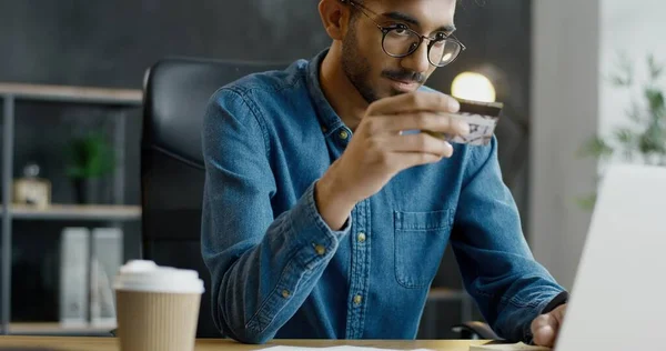 Arabiska unga manliga kontorsarbetare i glasögon sitter vid bordet, innehar kreditkort och handla online på bärbar dator. — Stockfoto