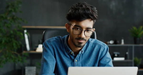 Närbild av snygga unga arabiska upptagen manliga kontorsarbetare sitter vid bordet och arbetar på bärbar dator. — Stockfoto