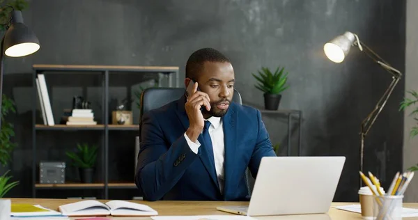 African American manliga kontorsarbetare sitter vid bordet, prata på smartphone medan du arbetar på bärbar dator. — Stockfoto