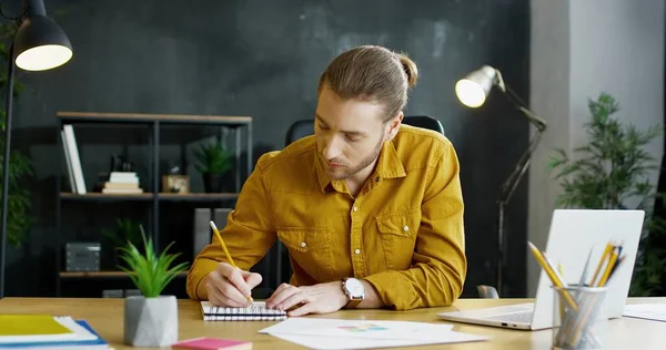 Ung affärsman sitter vid bordet på kontoret och skriver i anteckningar i anteckningsbok, arbetar på bärbar dator. — Stockfoto