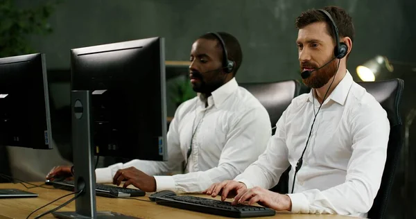 Blandade raser manliga call center medarbetare i trådlösa headset sitter vid skrivbordet framför datorskärmen och ger stöd till kunder. — Stockfoto