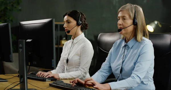 Zwei Callcenter-Mitarbeiterinnen sitzen am Tisch, tippen auf Tastaturen von Computern und unterhalten sich mit Kunden im Headset. — Stockfoto