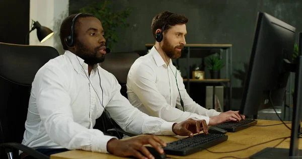 两个年轻英俊的多种族操作员，他们戴着耳机，在电脑上工作，与客户交谈. — 图库照片