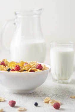 Bir kase mısır gevreği, sütlü, taze ahududu, yabanmersini. Sağlıklı kahvaltı için bir sürahi süt ve beyaz tahta üzerinde bir bardak.