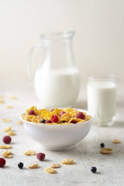 Bir kase mısır gevreği, sütlü, taze ahududu, yabanmersini. Sağlıklı kahvaltı için bir sürahi süt ve beyaz tahta üzerinde bir bardak.