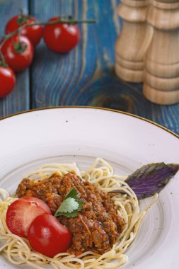 Beyaz tabakta spagetti bolonez makarna, tuz, biber, peynir ve domates mavi ahşap arka planda.