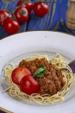Beyaz tabakta spagetti bolonez makarna, tuz, biber, peynir ve domates mavi ahşap arka planda.