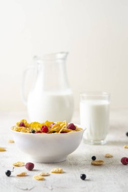 Bir kase mısır gevreği, sütlü, taze ahududu, yabanmersini. Sağlıklı kahvaltı için bir sürahi süt ve beyaz tahta üzerinde bir bardak.